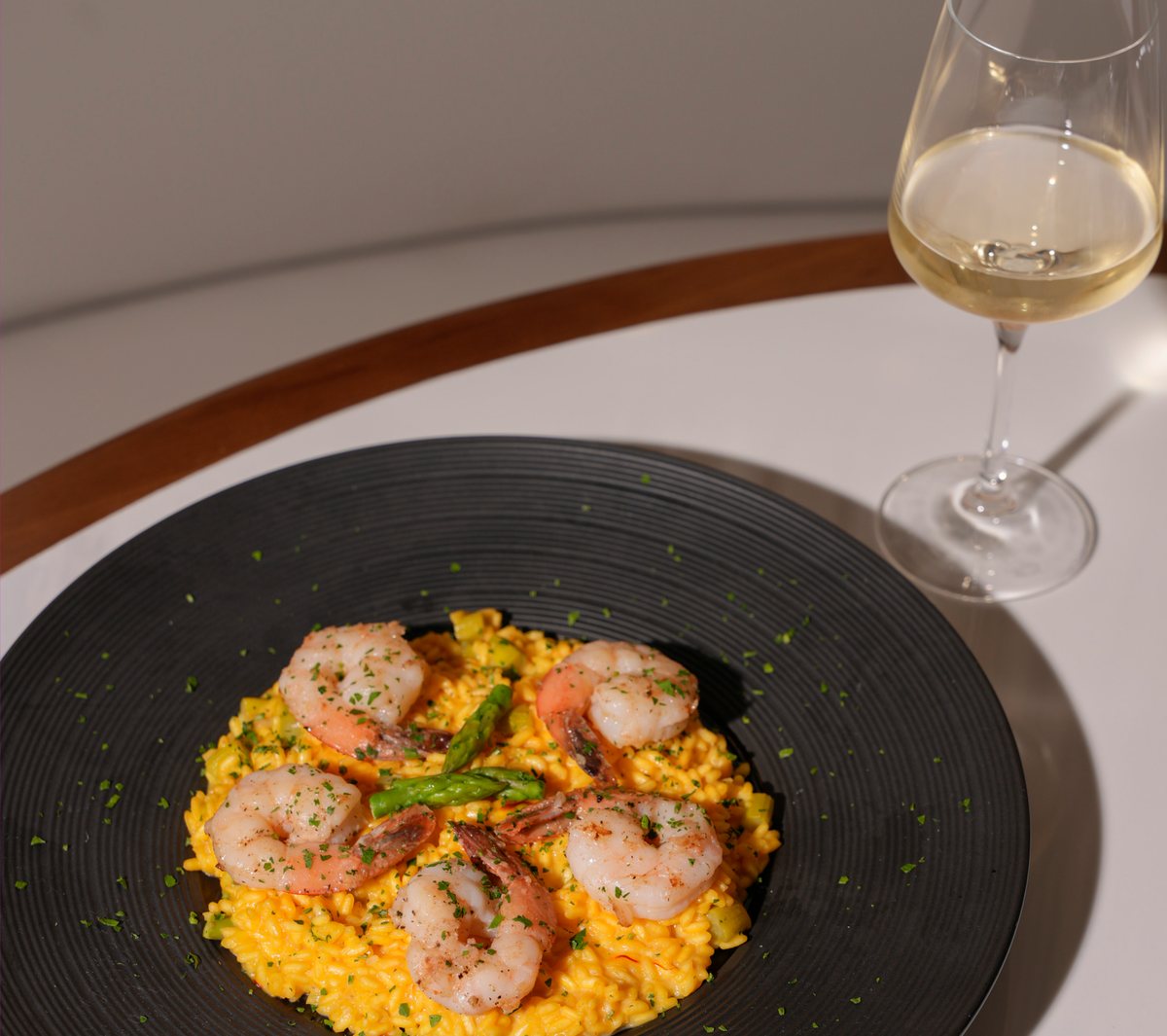Risotto allo zafferano con gamberi scottati e asparagi su un piatto nero, accanto un calice di bianco